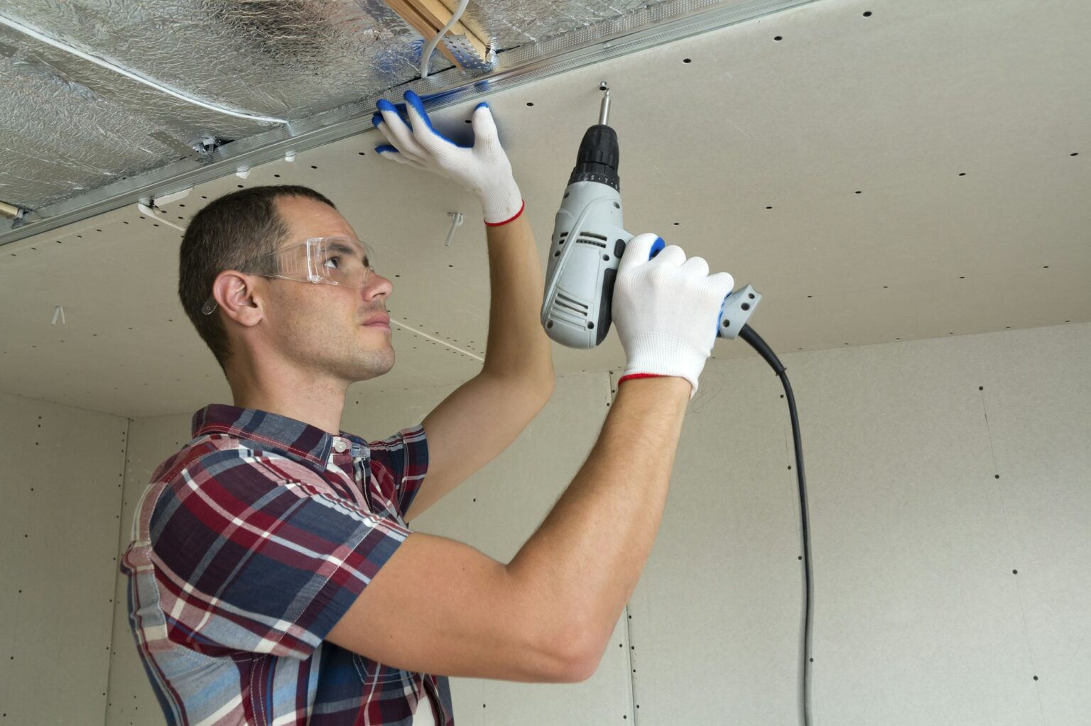 Young man in goggles fixing drywall suspended ceiling to metal frame using electrical screwdriver